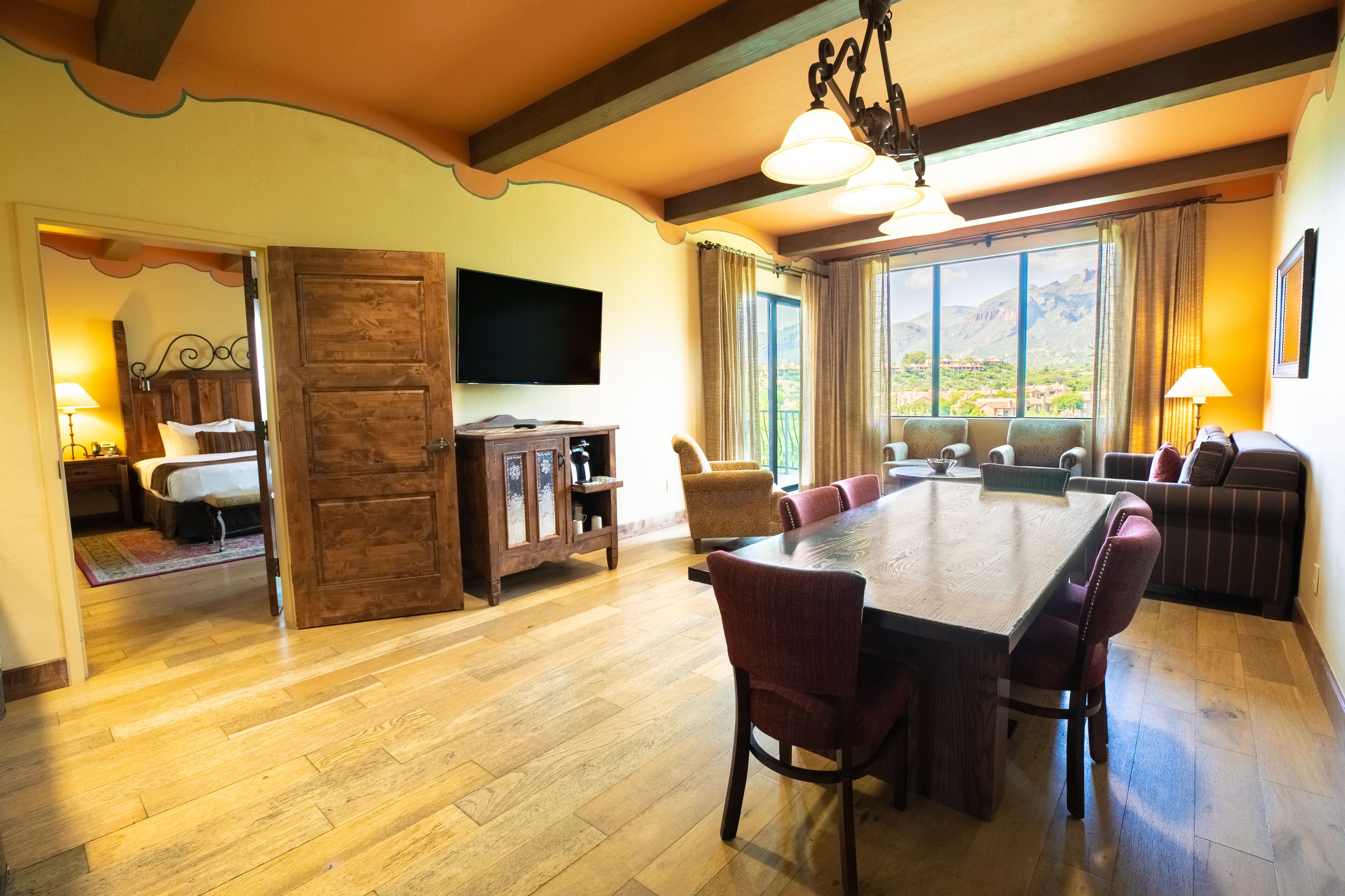 A spacious dining area with a large wooden table and multiple chairs, a television mounted on the wall, and a view of mountains through large windows.
