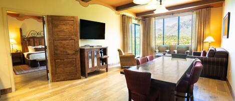 A spacious dining area with a large wooden table and multiple chairs, a television mounted on the wall, and a view of mountains through large windows.