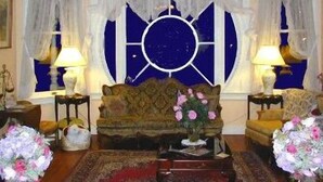 Una habitación con un sofá, dos mesitas auxiliares, un ventanal decorado con cortinas y un arreglo floral.