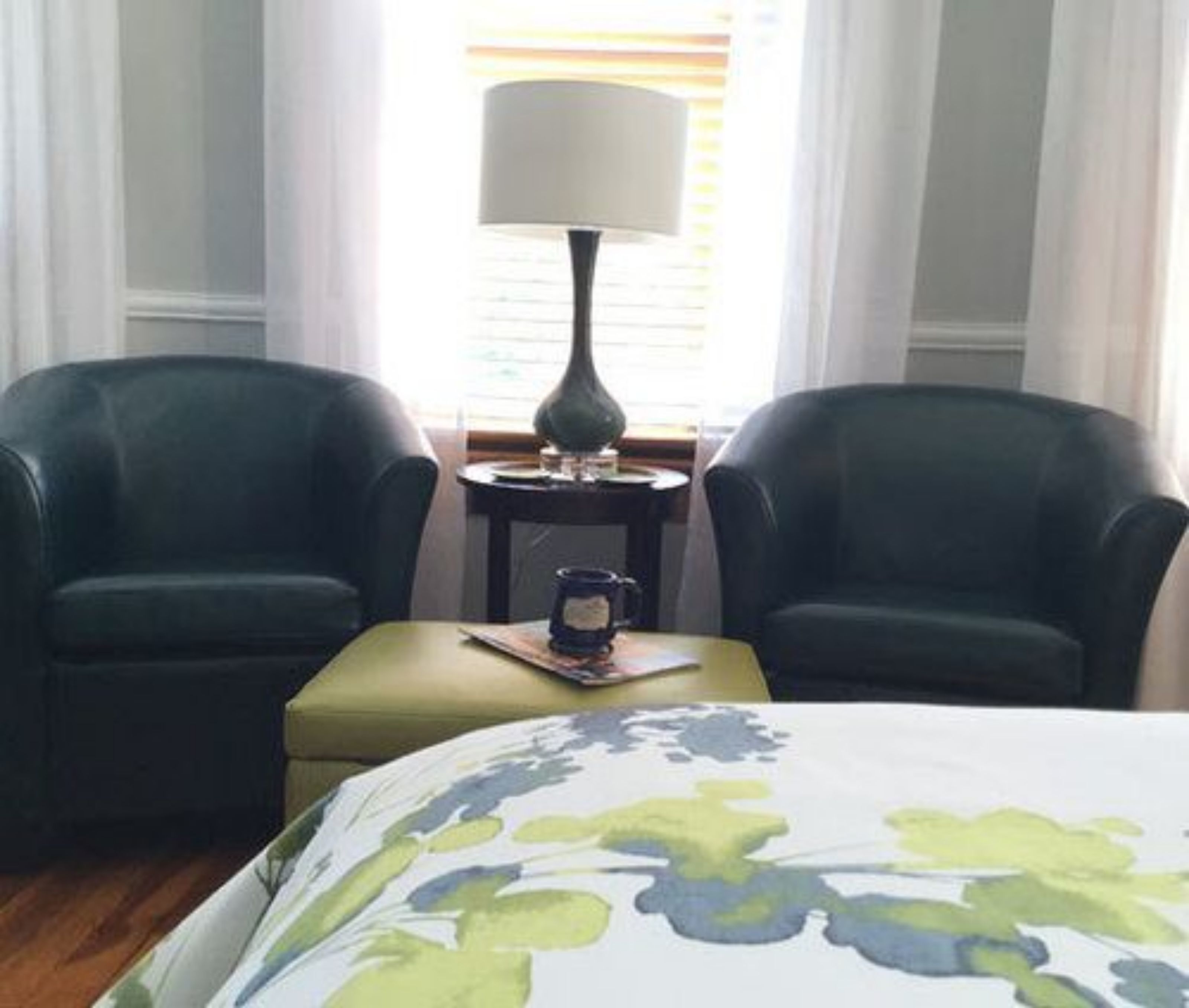 Une chambre d’hôtel avec deux fauteuils vert foncé, une petite table ronde avec une lampe et un lit recouvert d’une couvre-lit à motifs floraux.