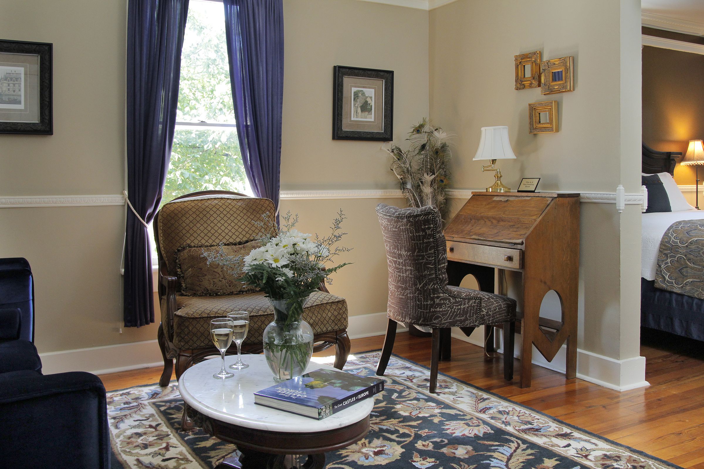 A room with a bed, a desk, a chair, a table with a vase and a book, and a window with curtains.