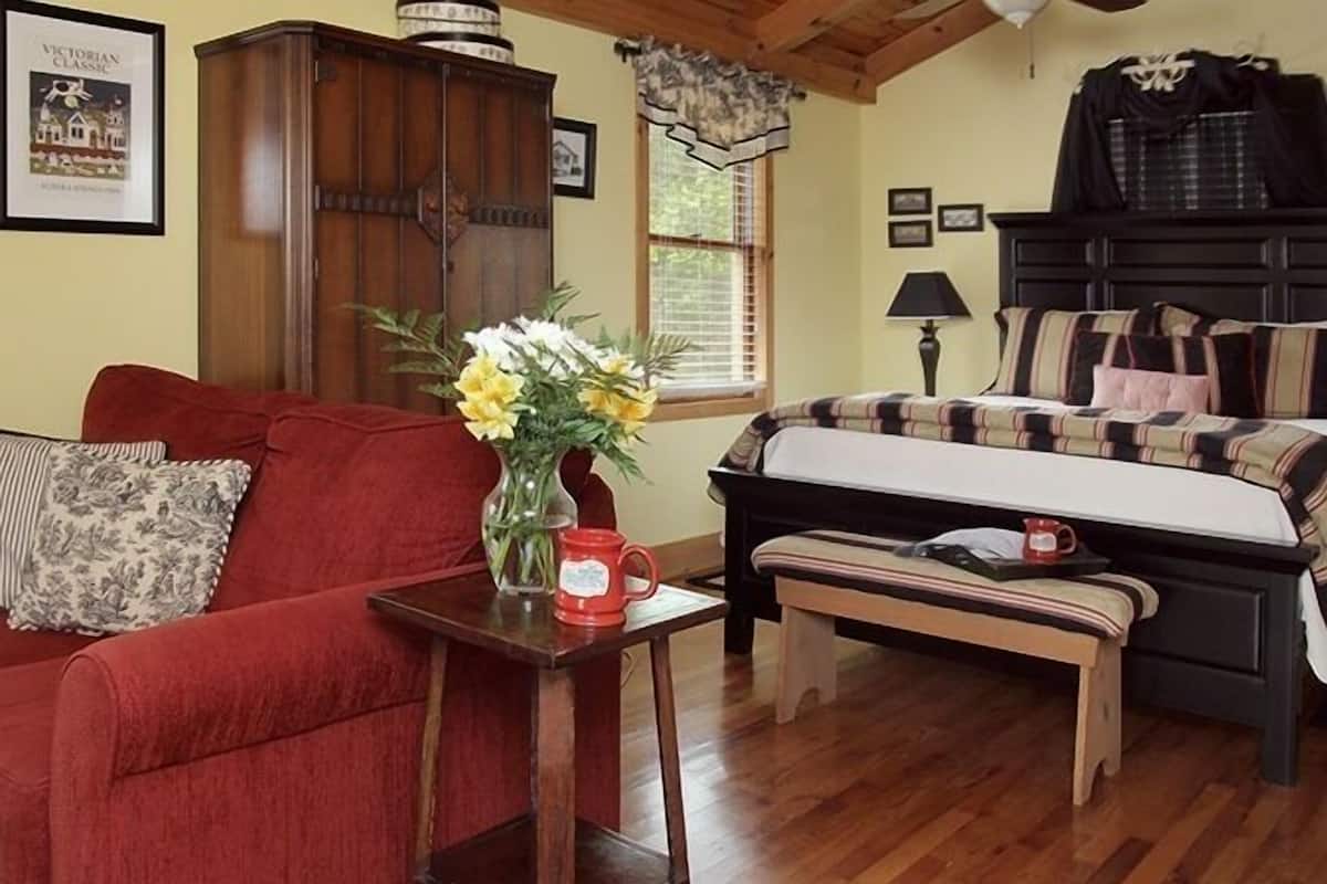 Een gezellige kamer met een rode bank, een houten kast, een bed met geruite beddengoed en een klein tafeltje met een bloemenvaas.