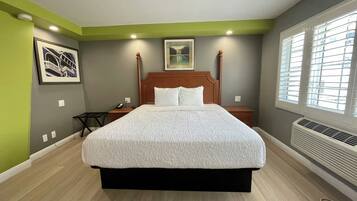 A neatly arranged bedroom with a large bed, a bedside table, a nightstand, a framed picture, and a window with blinds.