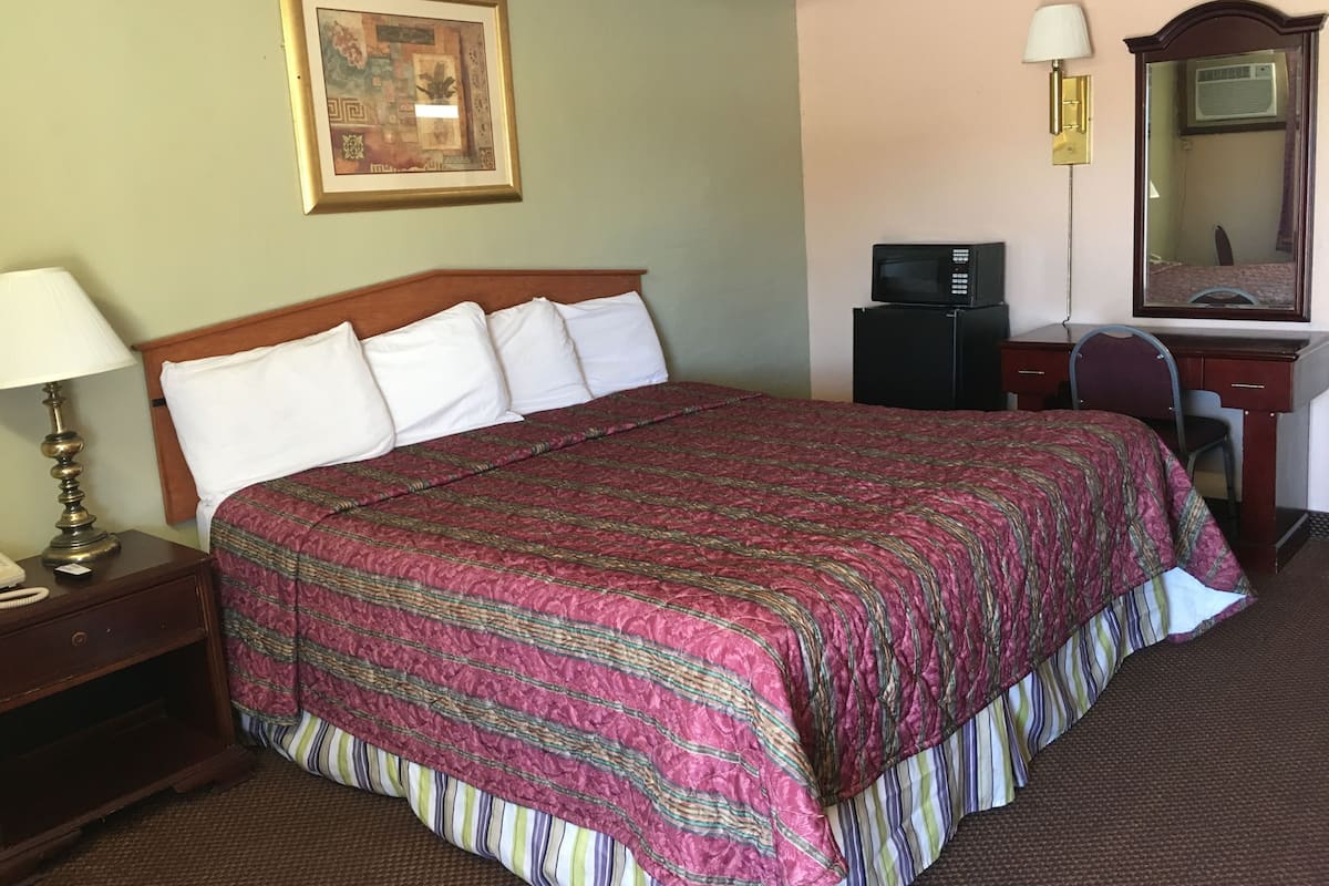 A neatly made bed with a patterned bedspread, a bedside lamp, a small desk with a computer, a mirror, and a framed picture on the wall.