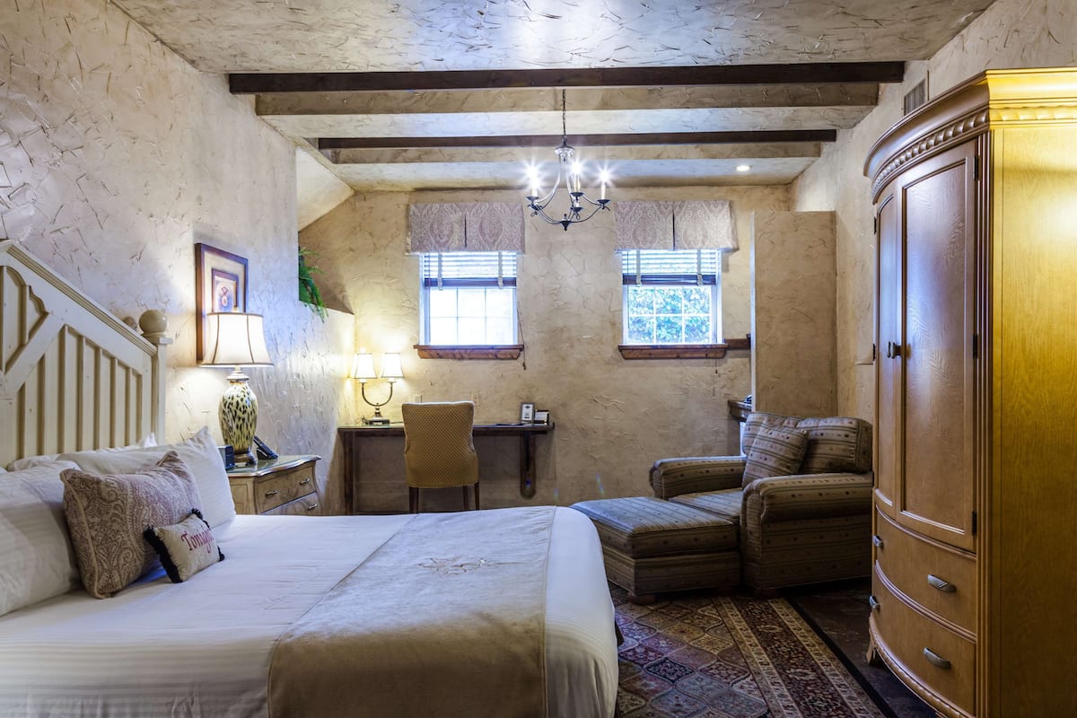A bedroom with a large bed, a wooden wardrobe, a desk with a chair, and two windows with blinds.