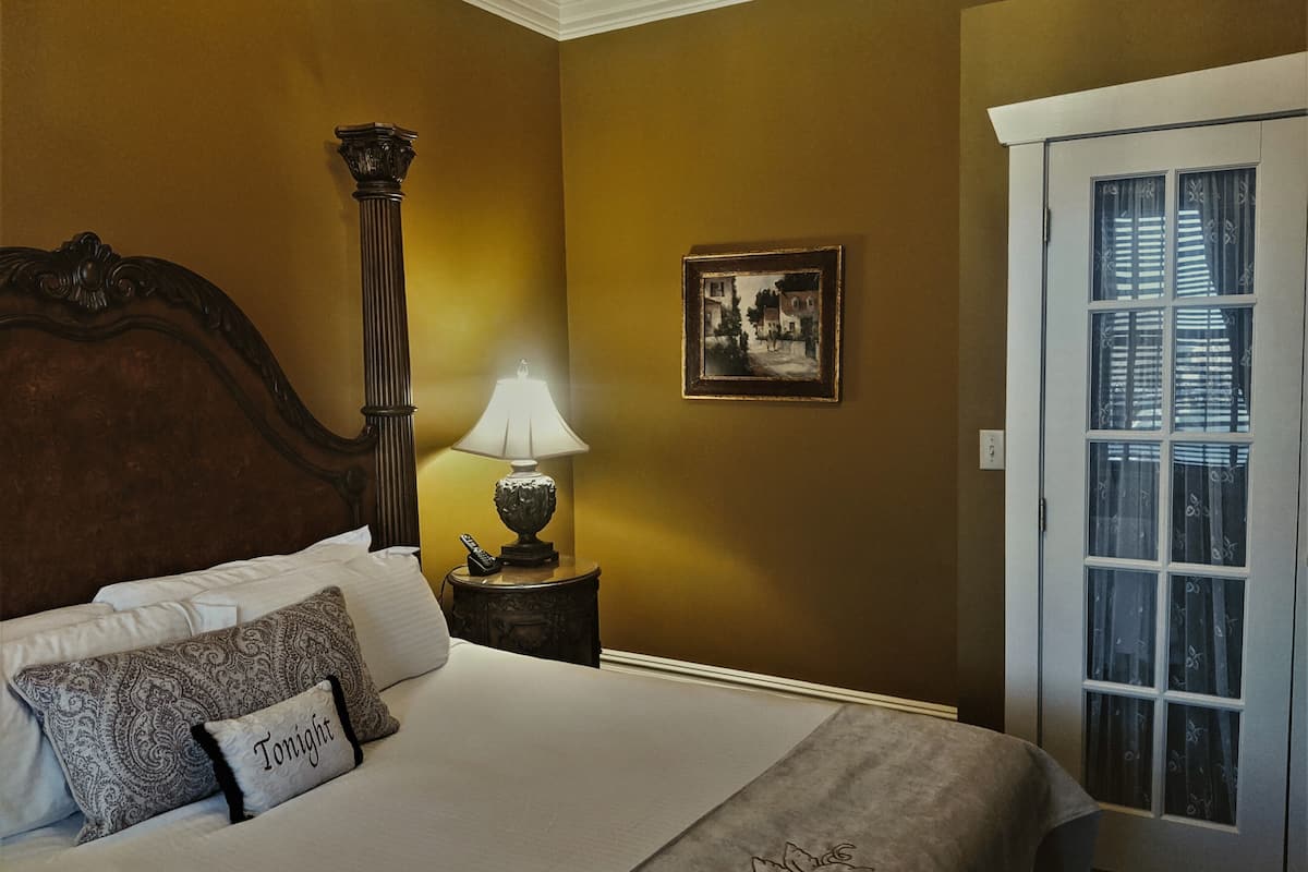 A bedroom with a large bed, a nightstand with a lamp, a framed picture on the wall, and a door with patterned glass.