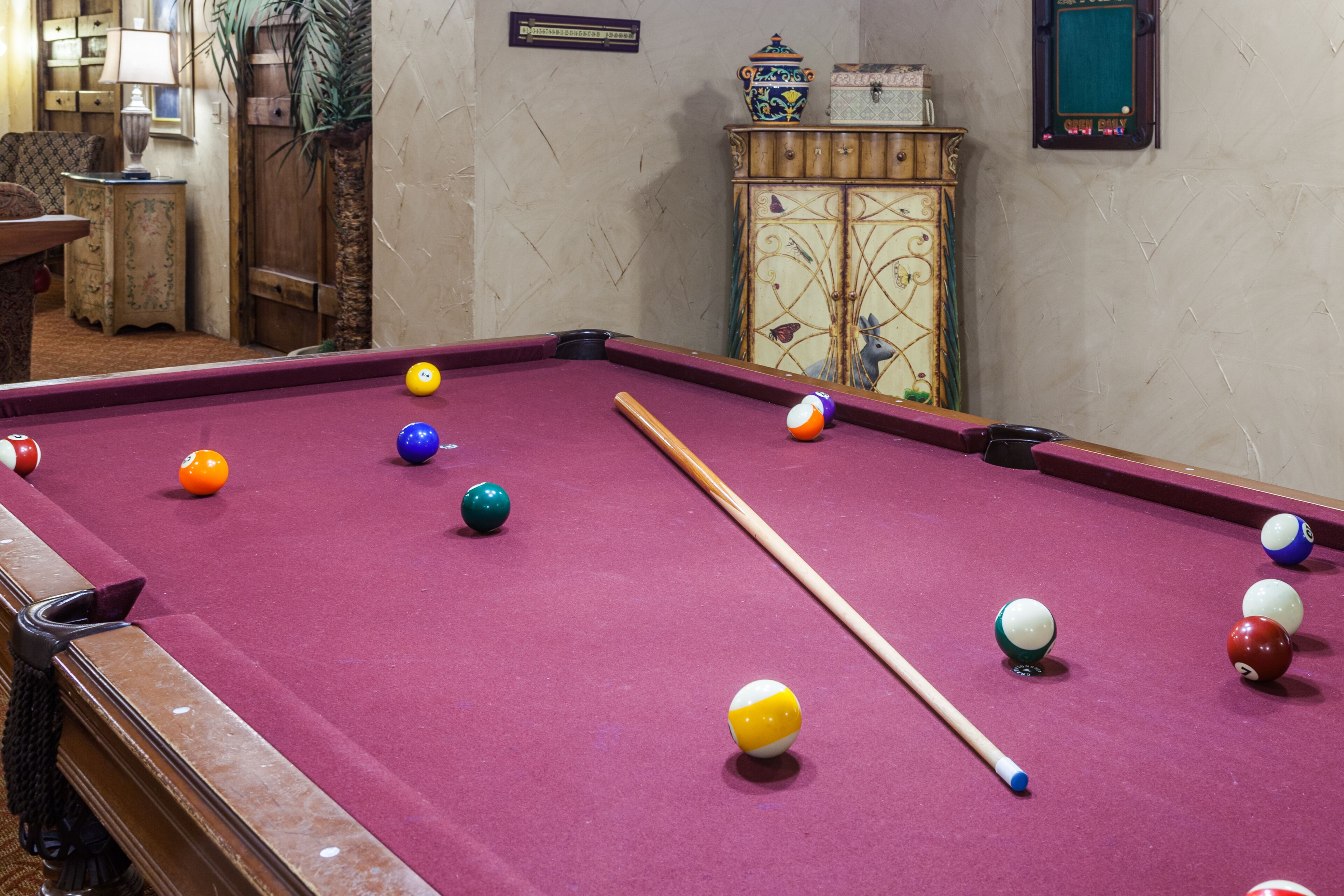 A pool table with balls and a cue stick, surrounded by a wooden cabinet and a lamp.
