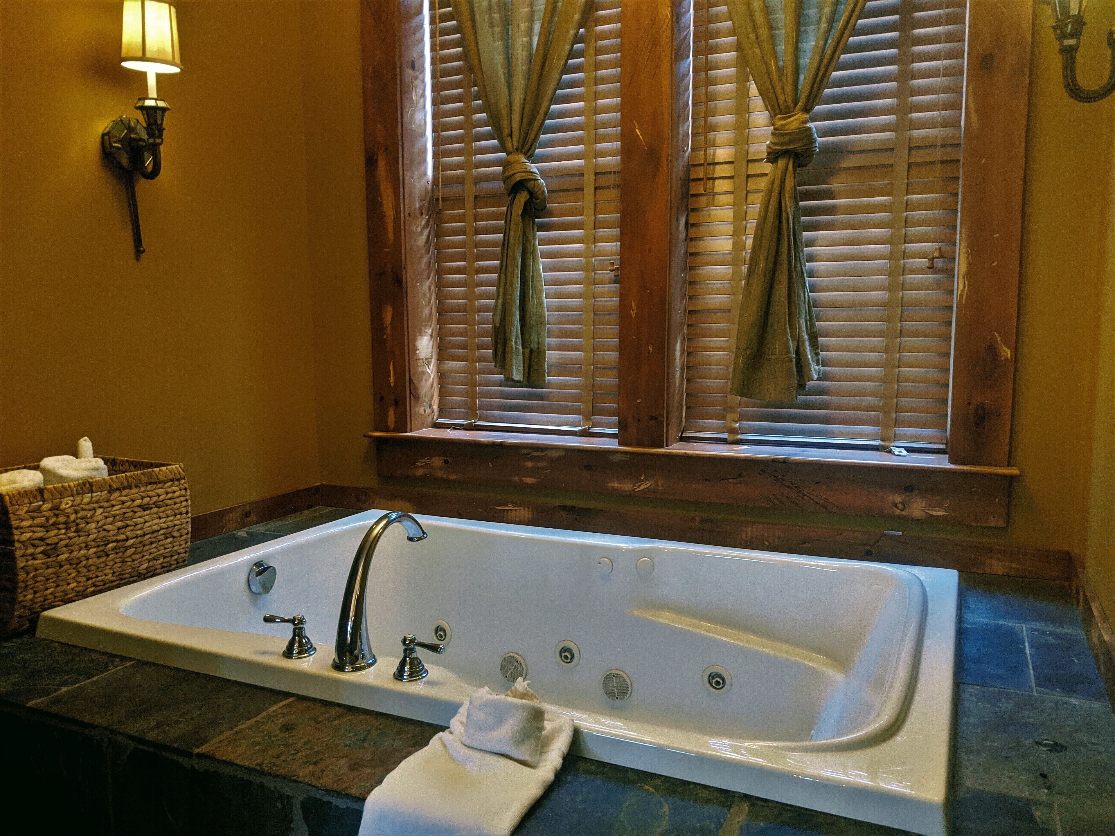 A bathroom with a large bathtub, a wooden window with curtains, a wall-mounted light fixture, and a wicker basket with towels.