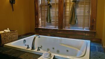 A bathroom with a large bathtub, a wooden window with curtains, a wall-mounted light fixture, and a wicker basket with towels.