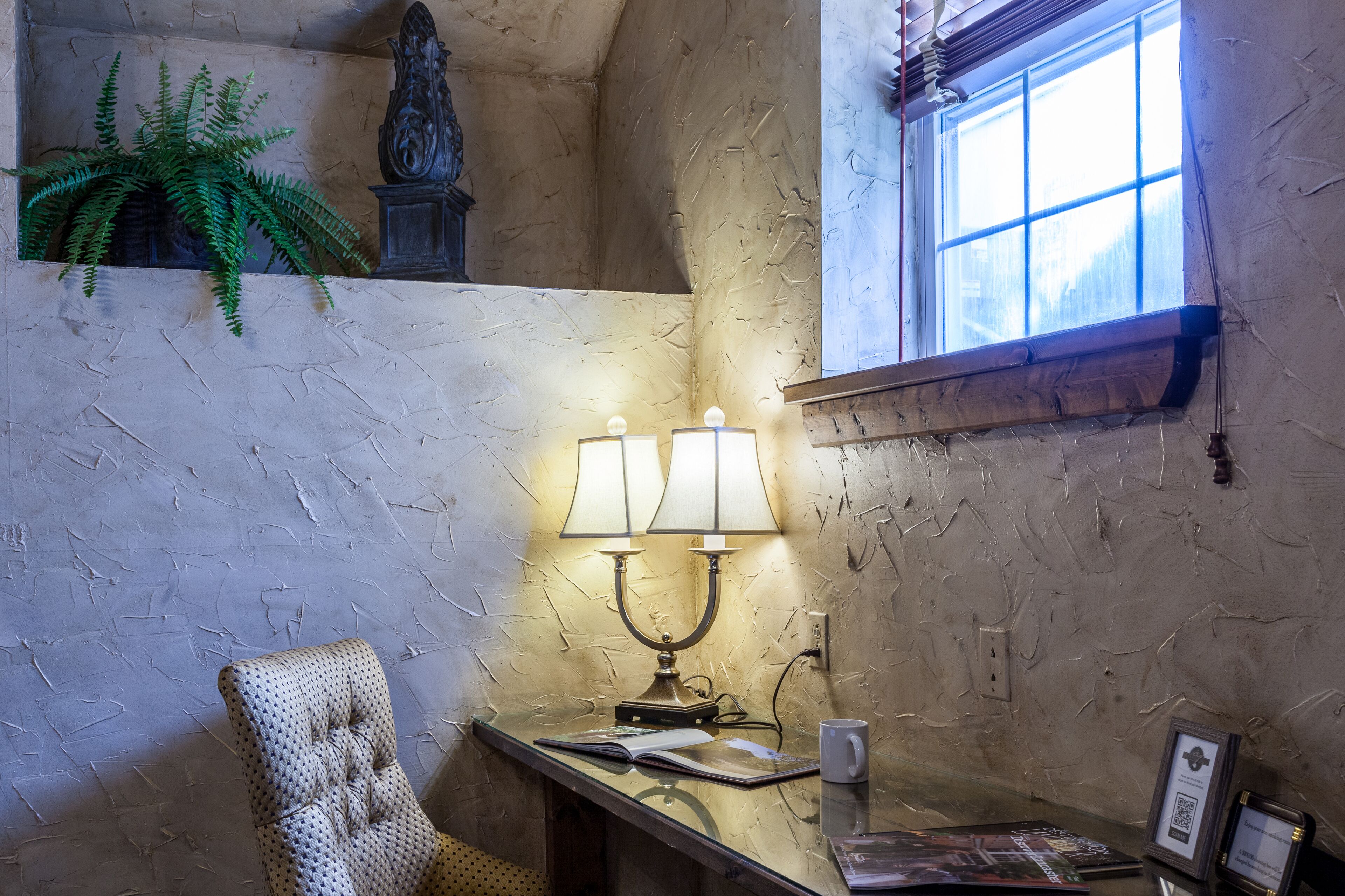 A room with a textured wall, a window with a view, a lamp, a chair, and a plant.