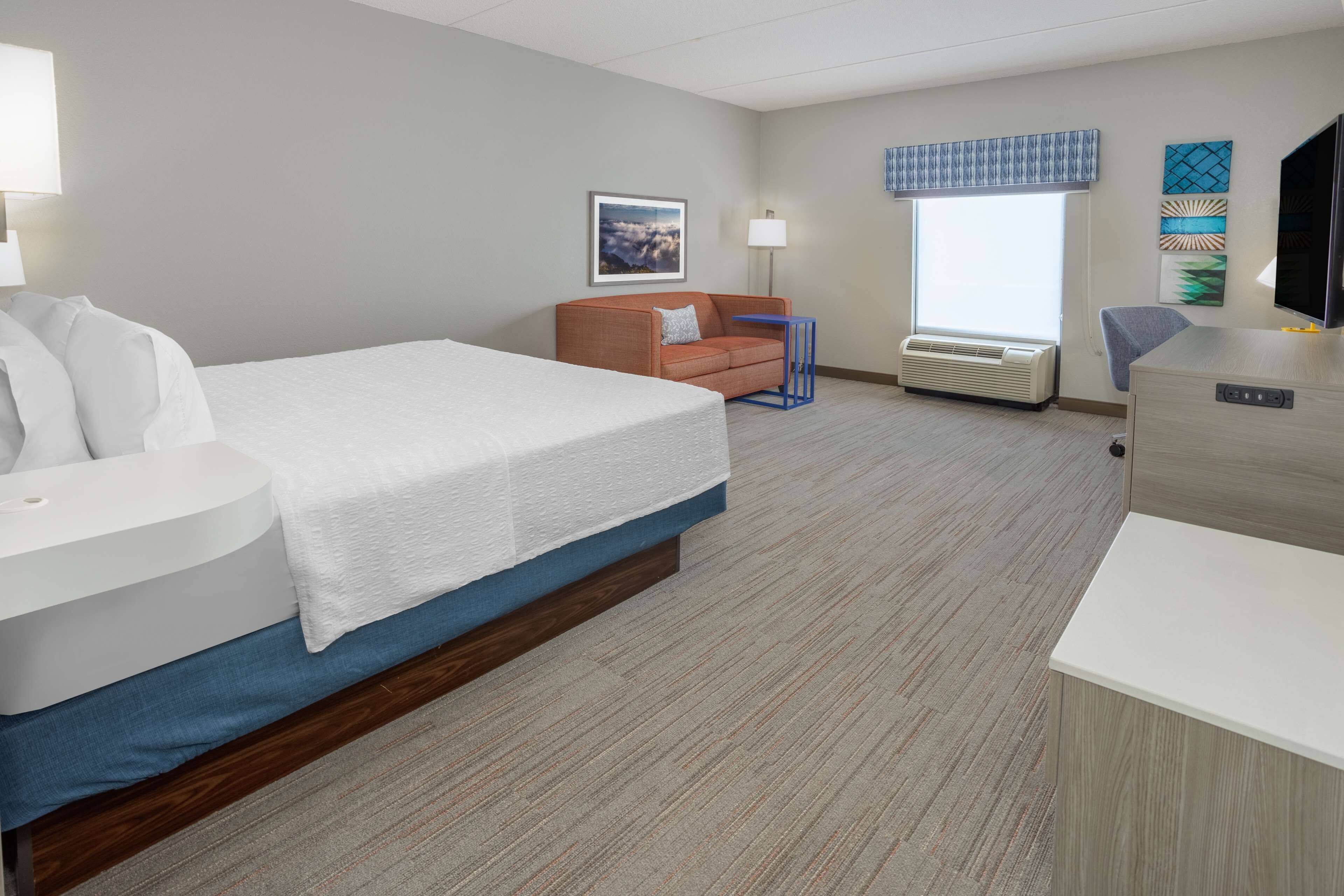 A hotel room with a bed, a desk, a chair, a sofa, a window with blinds, an air conditioner, and a wall-mounted TV.