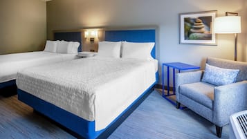Una camera d'albergo con due letti, una poltrona blu, un tavolino e una lampada a parete.
