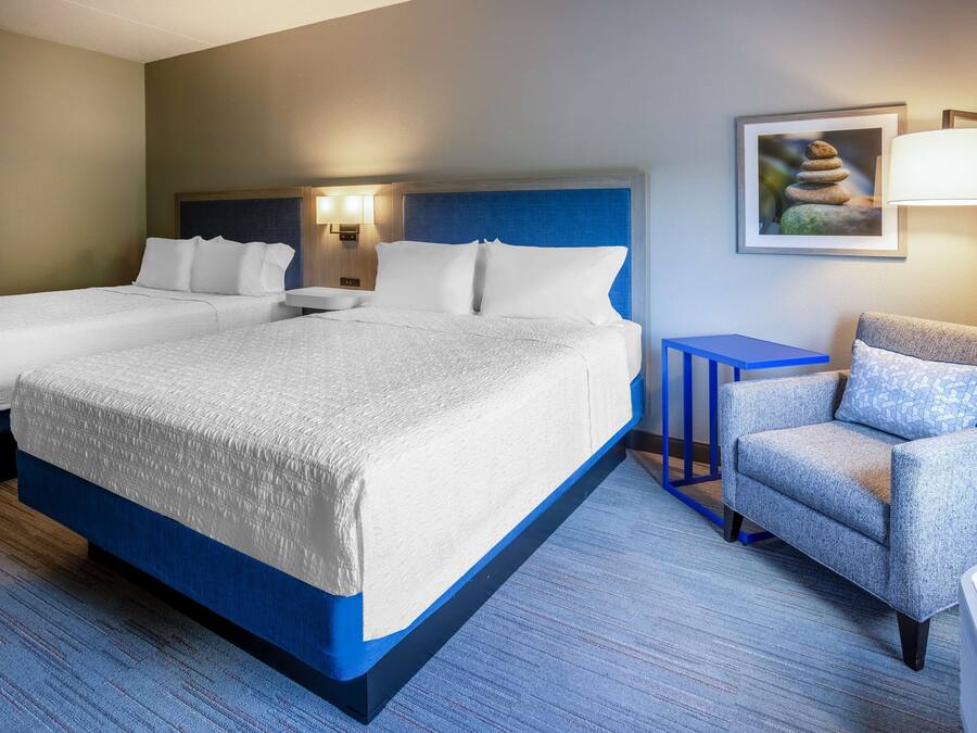 A hotel room with two beds, a blue armchair, a small table, and a wall-mounted lamp.