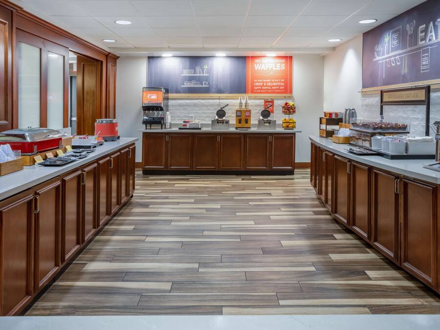 A hotel breakfast area with a buffet setup, including a coffee station, a waffle station, and various food items.
