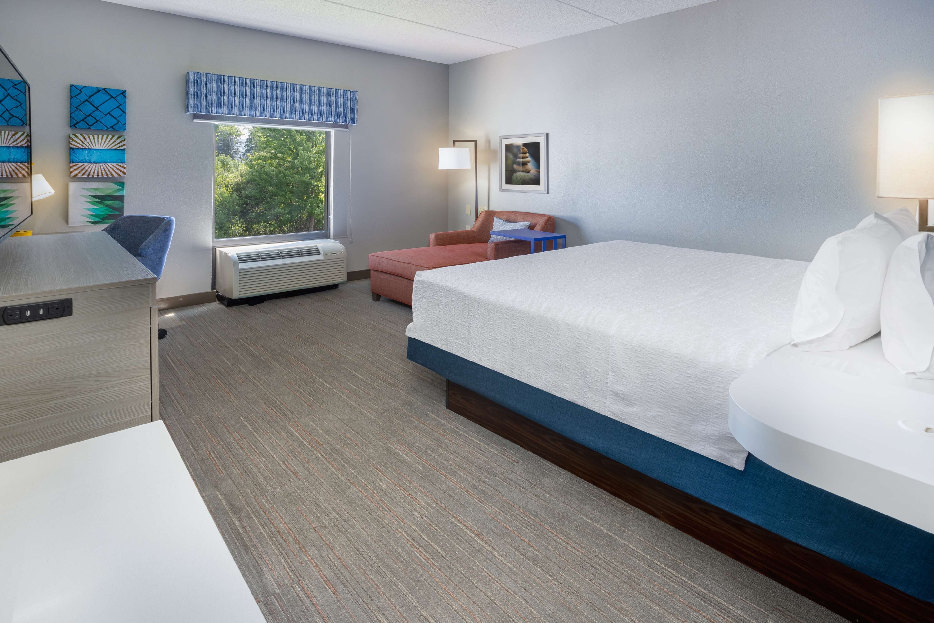 A hotel room with a large bed, a desk, a chair, and a window with a view of trees.