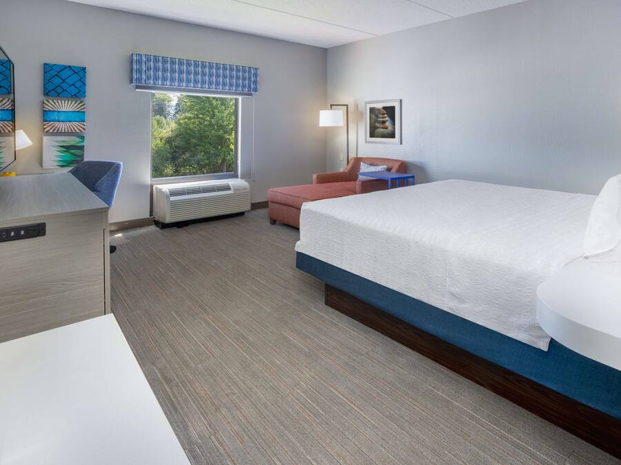 A hotel room with a large bed, a desk, a chair, and a window with a view of trees.