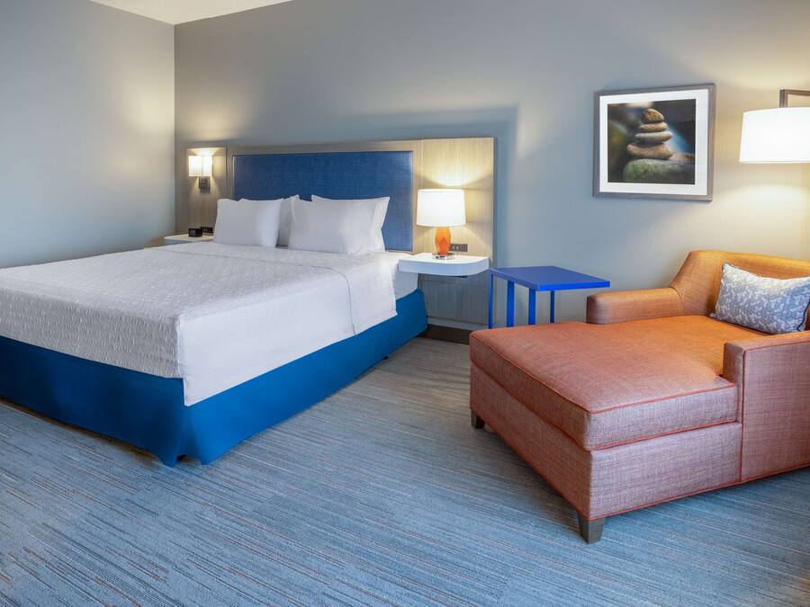 A hotel room with a large bed, a nightstand, a small table, an orange armchair, and a framed picture on the wall.