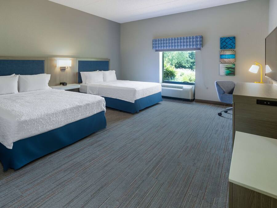 A hotel room with two beds, a desk, a chair, and a window with a view of greenery.