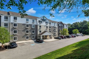 A multi-story hotel with a central entrance, parking lot, and surrounding greenery. - WoodSpring Suites Louisville Southeast (Louisville)