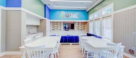 A dining area with white tables and chairs, a blue accent wall, and a kitchen area with white cabinets.