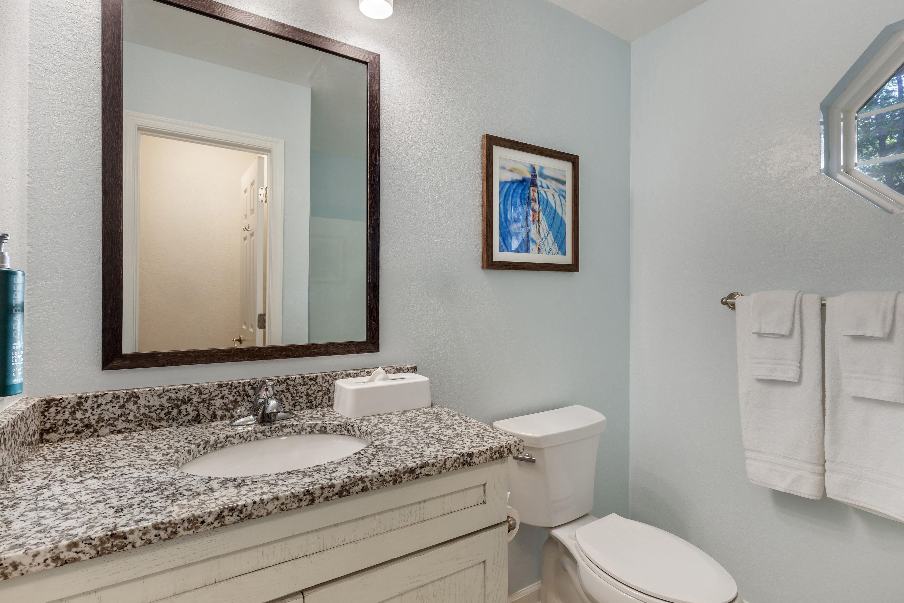 A bathroom with a granite countertop, a white sink, a toilet, a framed picture on the wall, and a towel rack with towels.