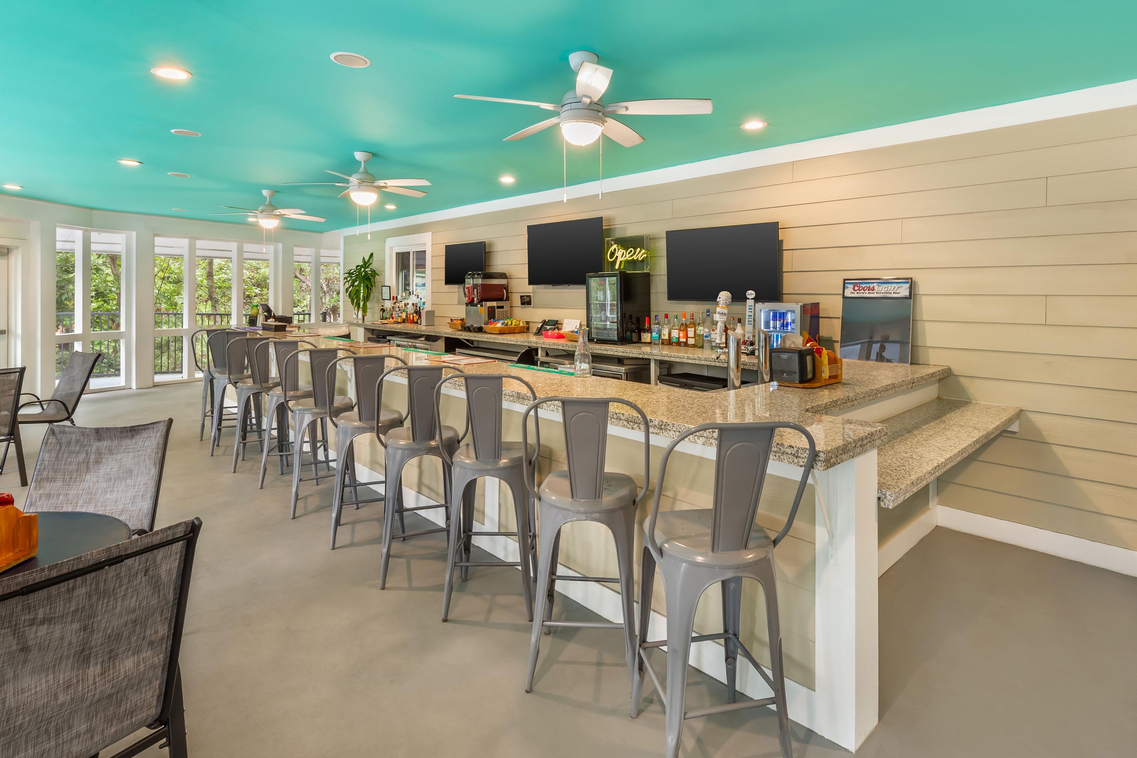 A bar area with a counter, bar stools, ceiling fans, and a large window with a view of trees.