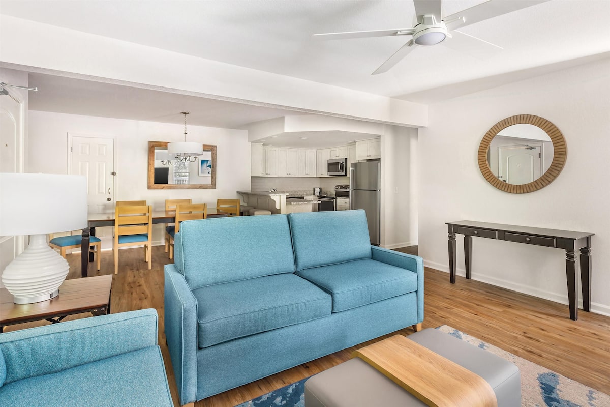 A modern living room with a blue sofa, wooden dining table, and a ceiling fan.