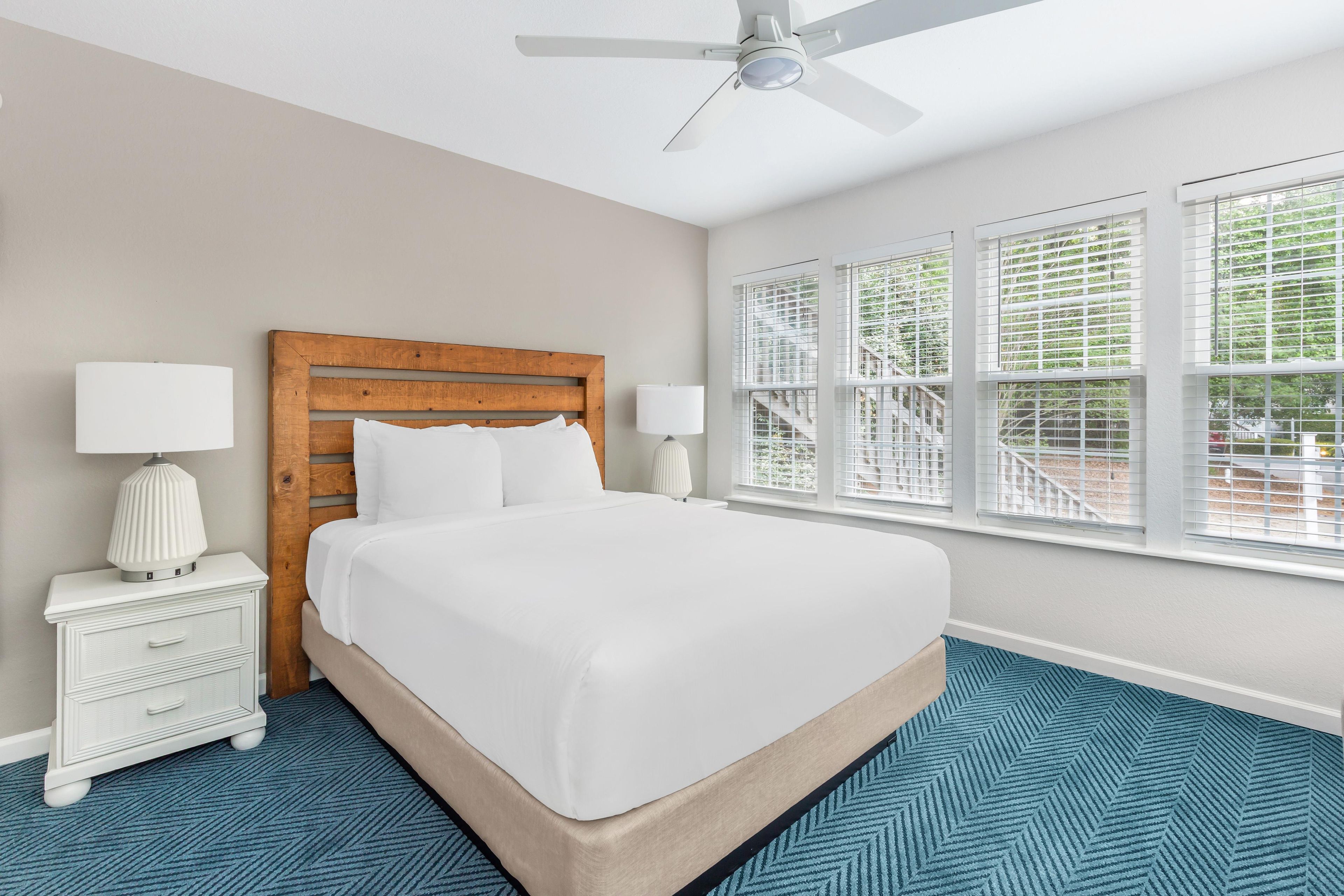 A hotel room with a large bed, two bedside tables with lamps, a ceiling fan, and large windows with blinds.