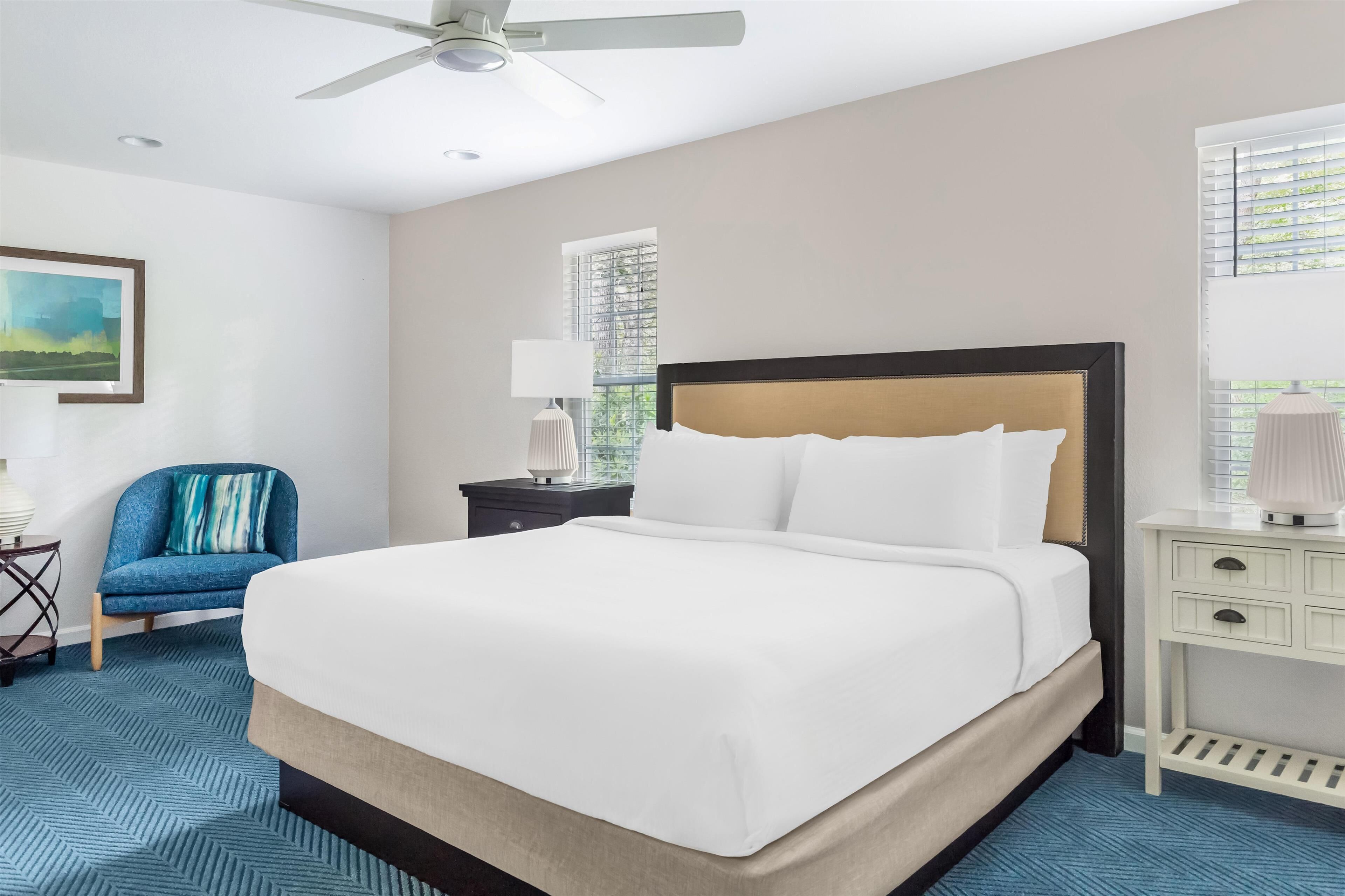 A hotel room with a large bed, a desk with a lamp, a chair, and a ceiling fan.