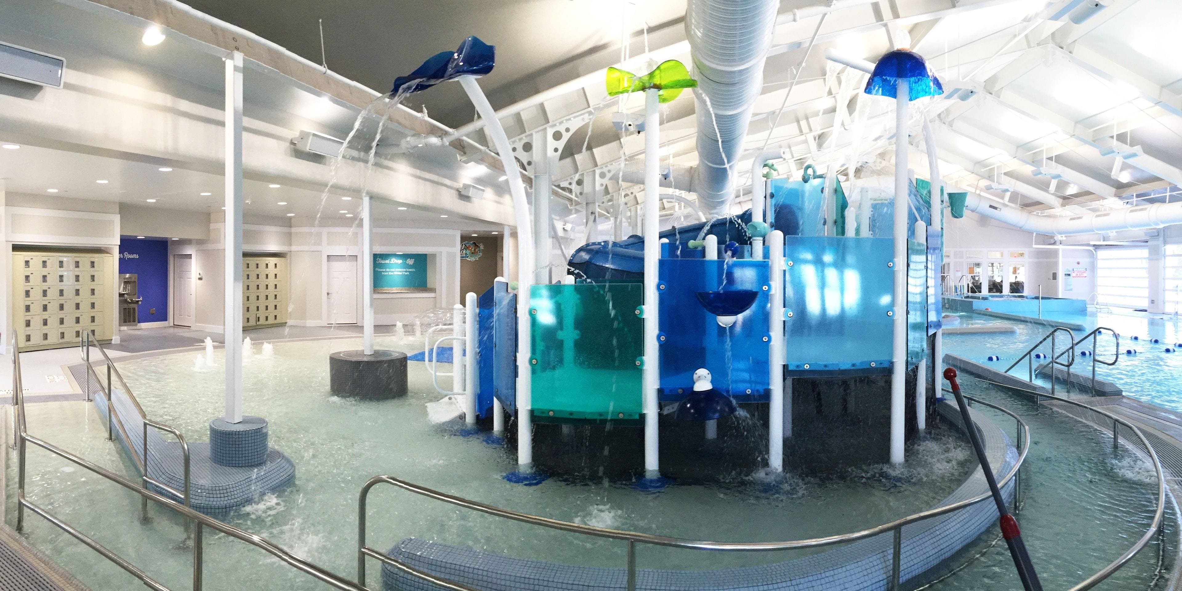 An indoor water feature with multiple fountains and water jets, surrounded by a pool area with a tiled floor and metal railings.