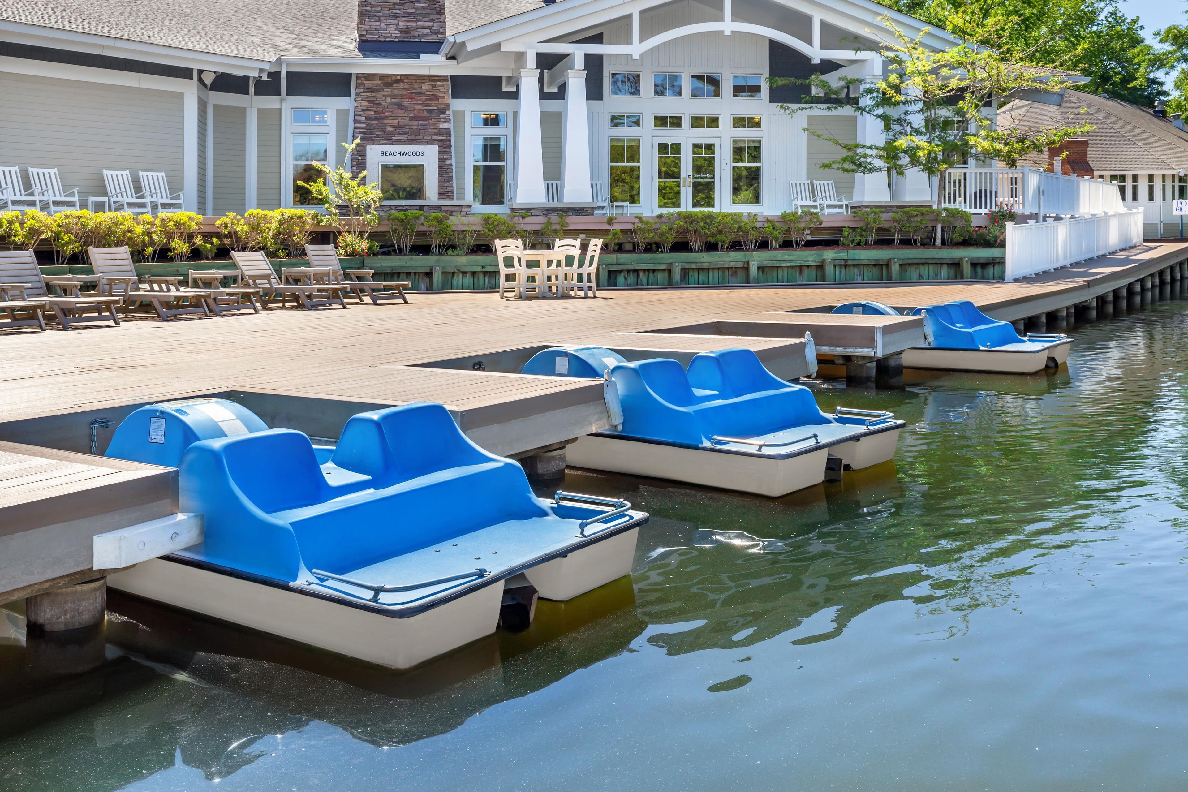 Une zone riveraine où sont amarrés des pédalos, un bâtiment portant une enseigne indiquant « BEACHWOODS », et des tables et chaises installées à l’extérieur.