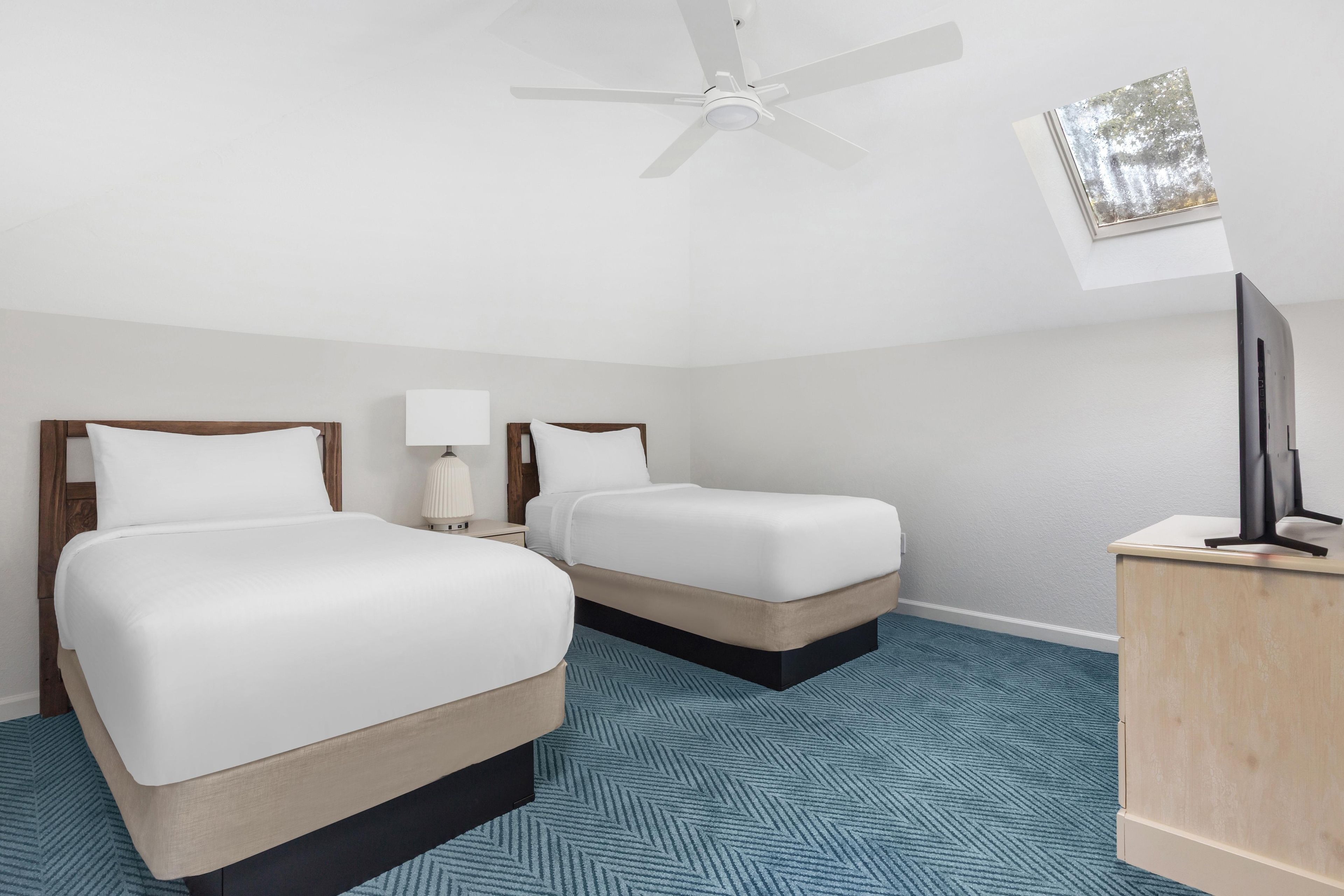 Habitación de hotel con dos camas, ventilador de techo, escritorio con lámpara y alfombra azul estampada.