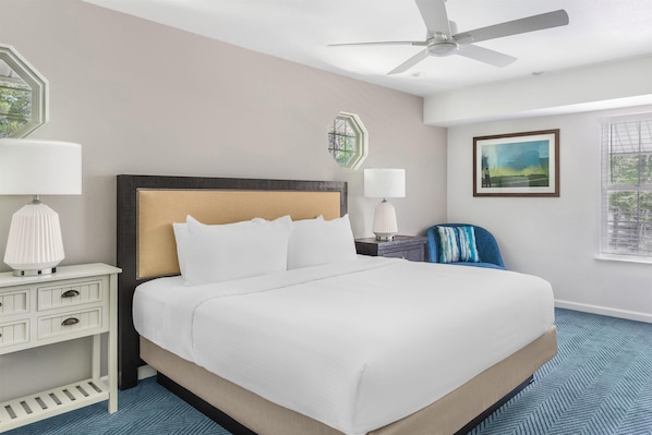 A hotel room with a large bed, a ceiling fan, a window with blinds, a painting on the wall, and a nightstand with a lamp.
