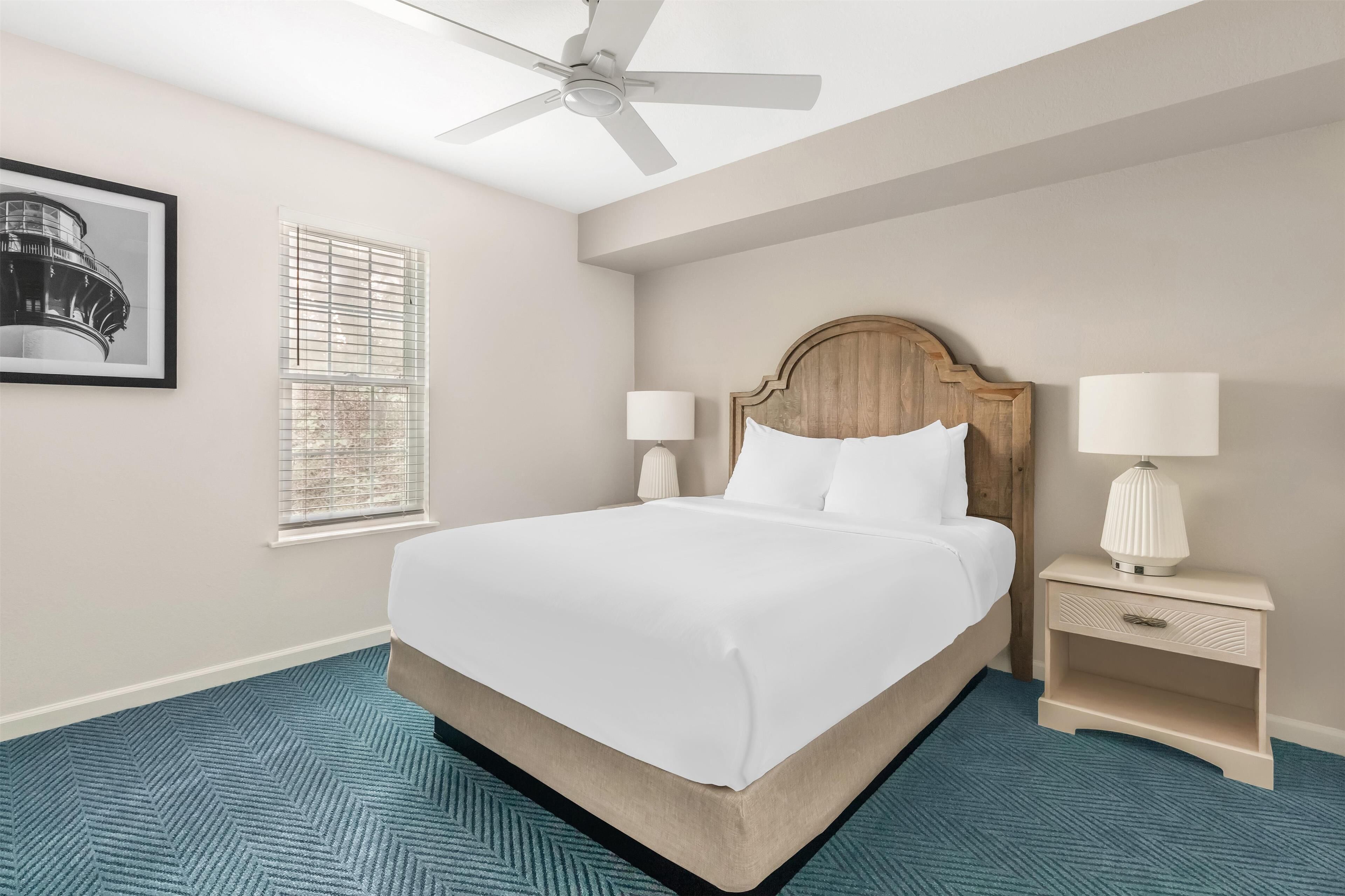 A hotel room with a large bed, two bedside lamps, a ceiling fan, a framed picture on the wall, and a window with blinds.