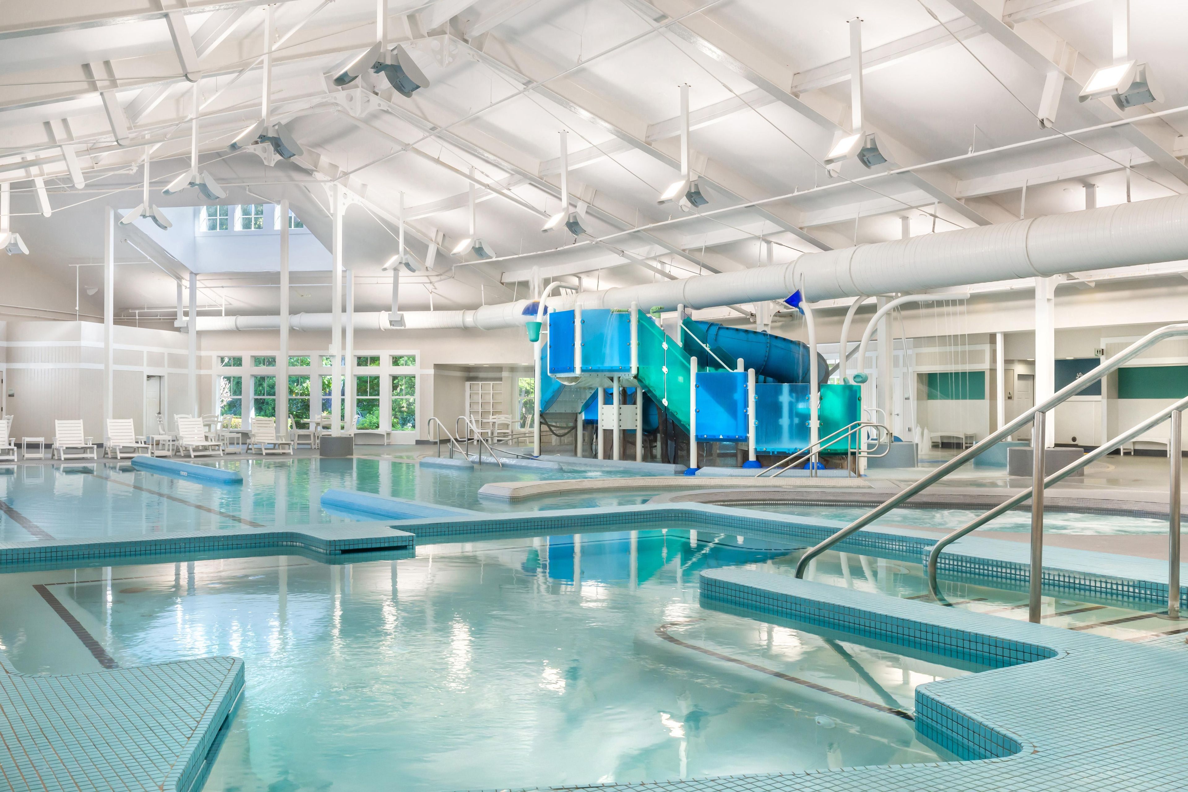 Une grande piscine intérieure dotée de plusieurs couloirs de nage, d’un toboggan et d’un espace détente avec des chaises.