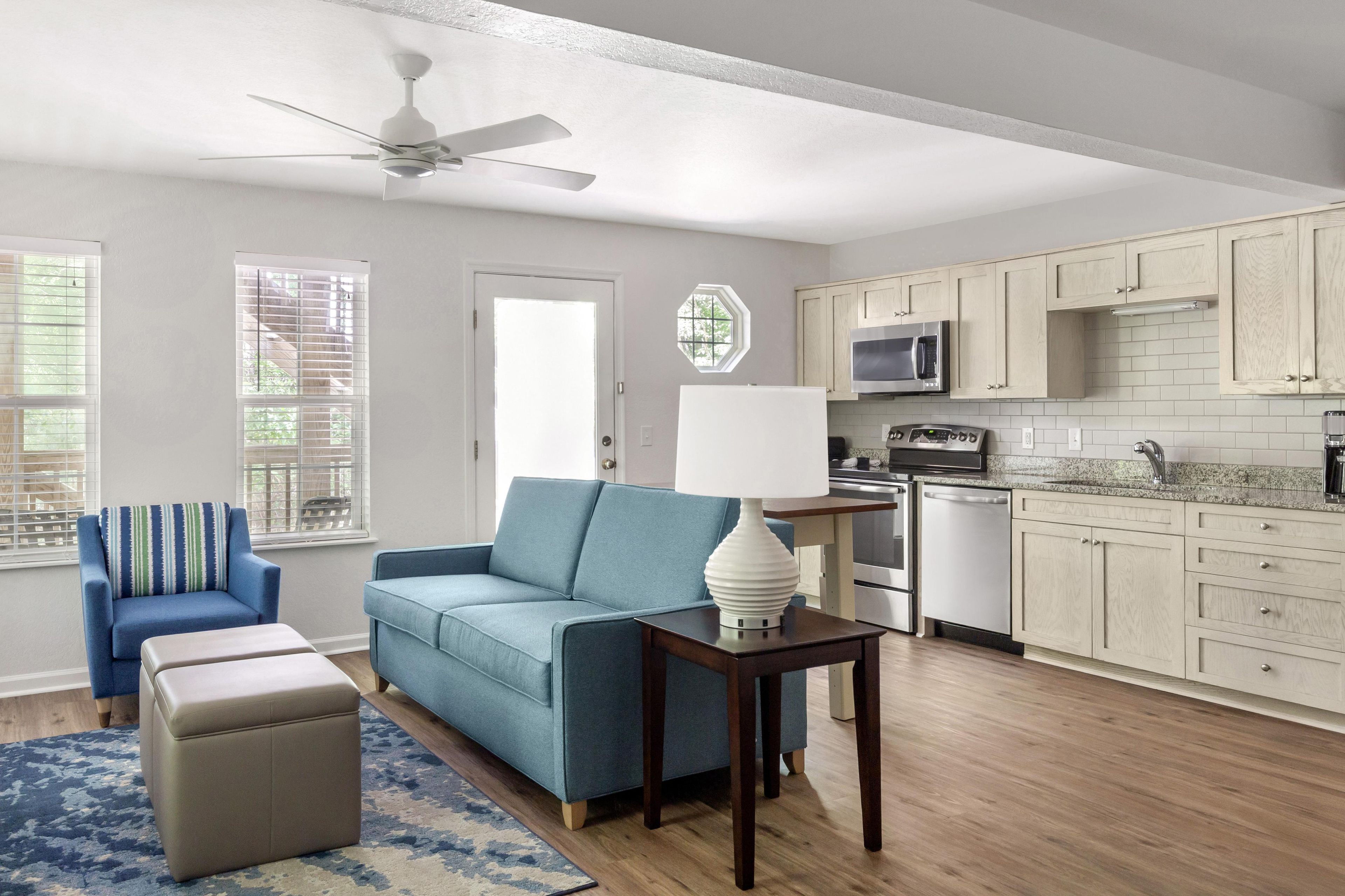 A modern living room with a sofa, ottoman, and a table with a lamp. There is a kitchen area with cabinets and appliances.