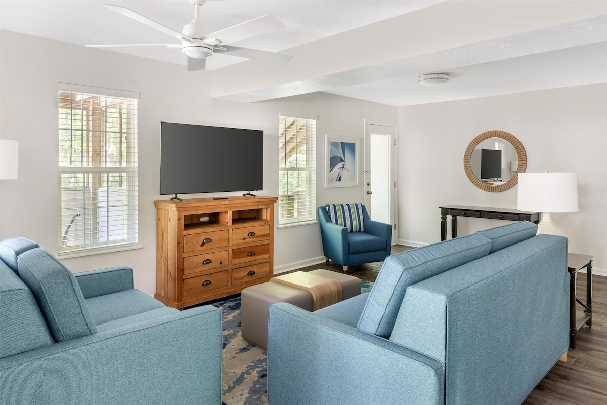 A living room with a ceiling fan, a flat-screen TV on a wooden cabinet, a blue sofa, a blue armchair, and a round mirror on a black table.