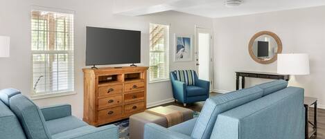 Una sala de estar con un ventilador de techo, un televisor de pantalla plana sobre un mueble de madera, un sofá azul, un sillón azul y un espejo redondo sobre una mesa negra.