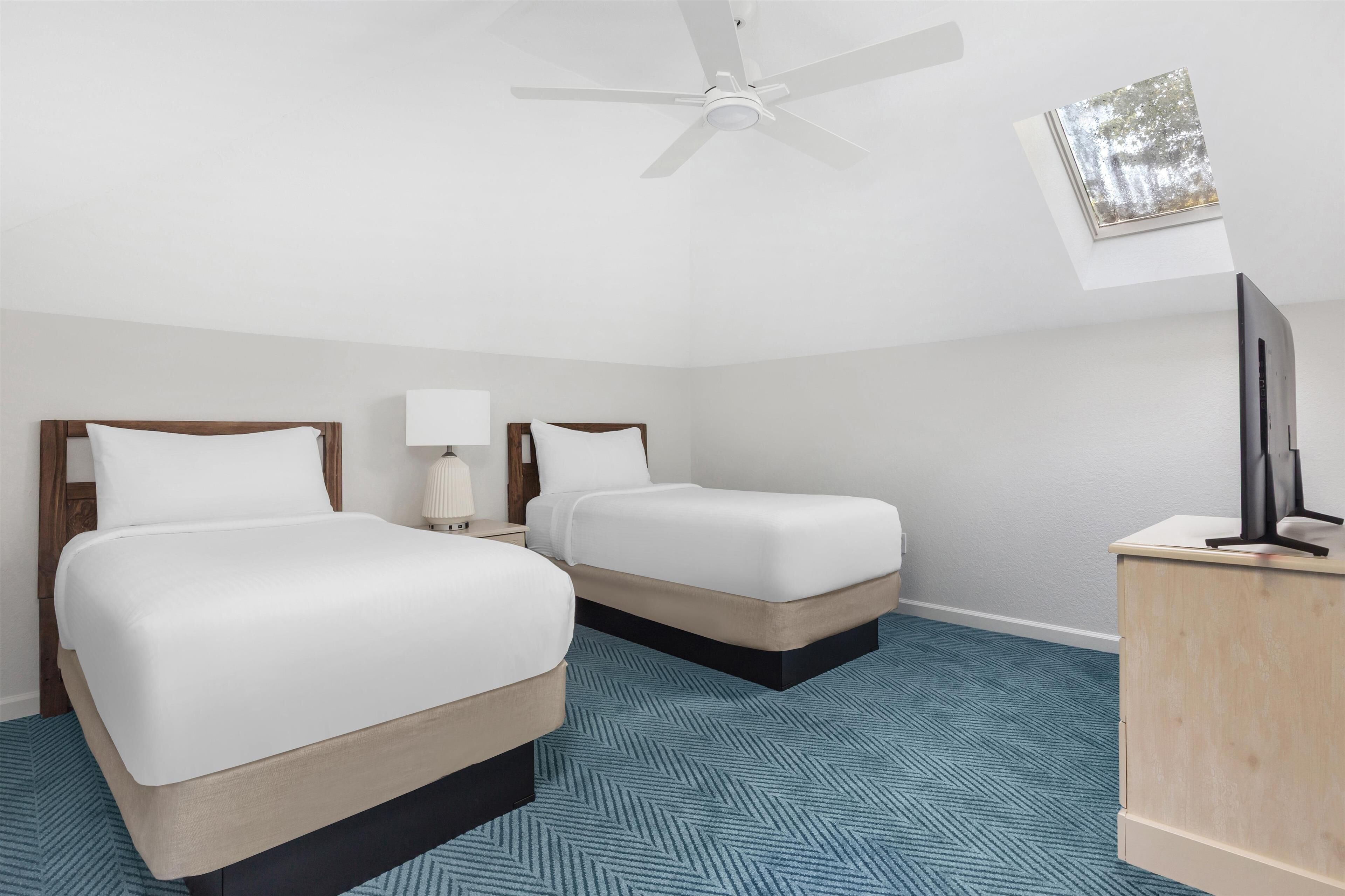 Une chambre d’hôtel avec deux lits, un ventilateur de plafond, un bureau avec une lampe et une petite fenêtre donnant sur un paysage agréable.