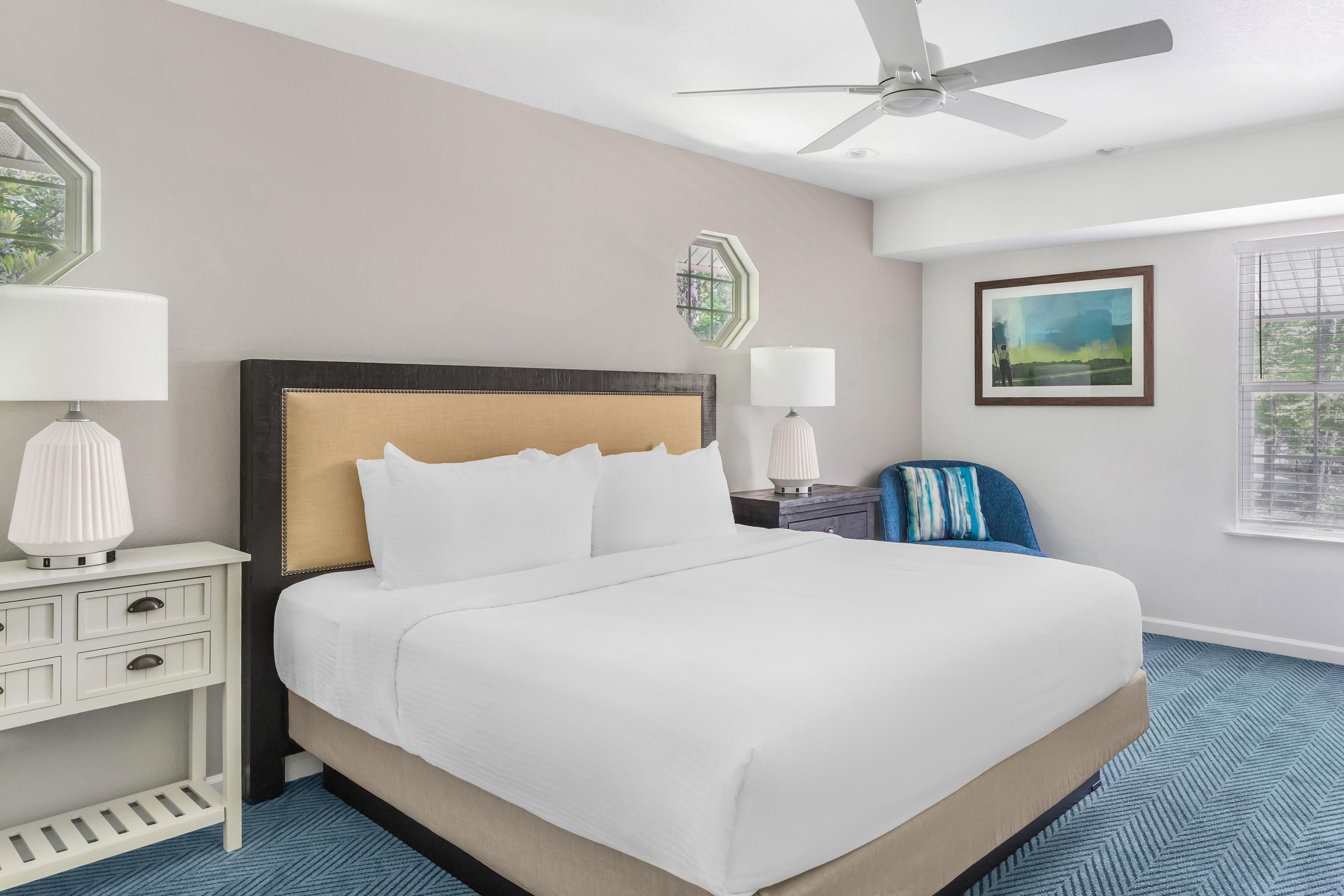 A hotel room with a bed, a nightstand, a lamp, a ceiling fan, a window with blinds, and a painting on the wall.
