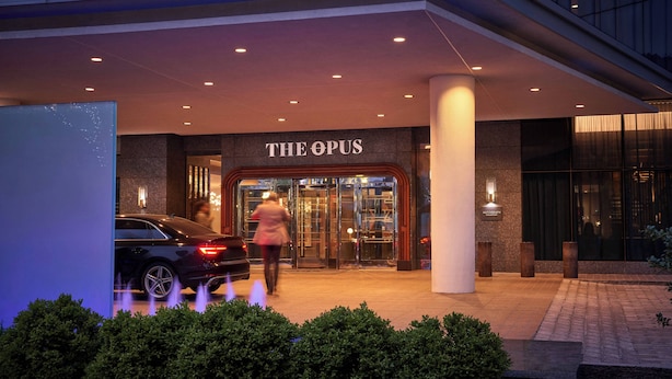 A modern hotel entrance with a glass door, a car parked outside, and a well-lit pathway leading to the entrance.