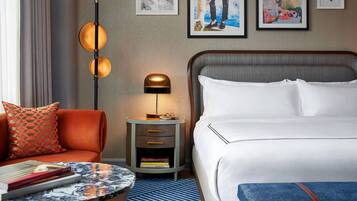 Quarto de hotel com uma cama, uma mesinha de cabeceira com abajur, uma mesa redonda com livros e um travesseiro estampado em vermelho.