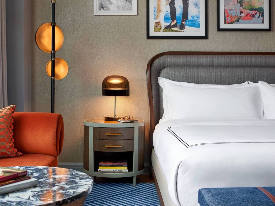 A hotel room with a bed, a nightstand with a lamp, a round table with books, and a red patterned pillow.