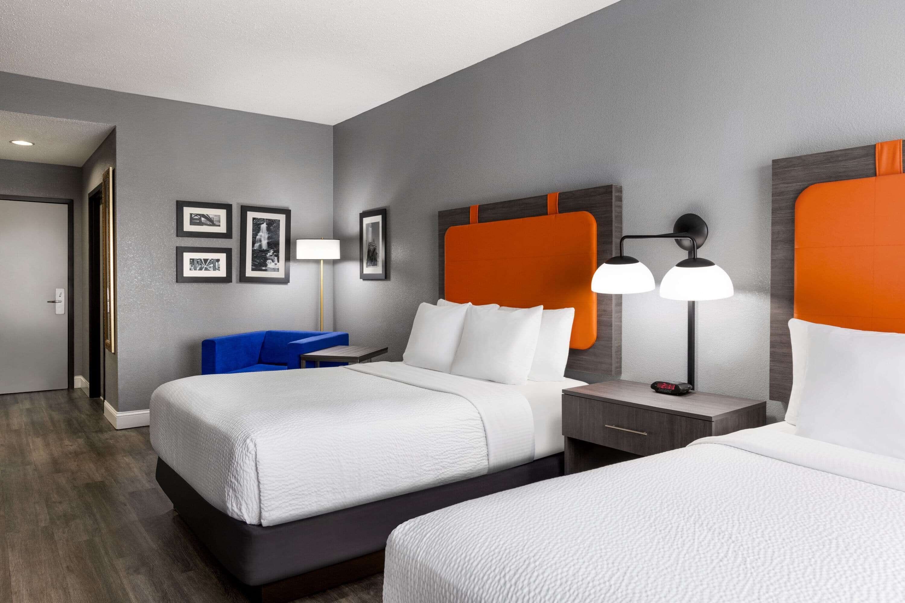 Quarto de hotel com uma cama grande, uma mesa de cabeceira, um abajur e quadros nas paredes. Há uma cadeira azul no canto.