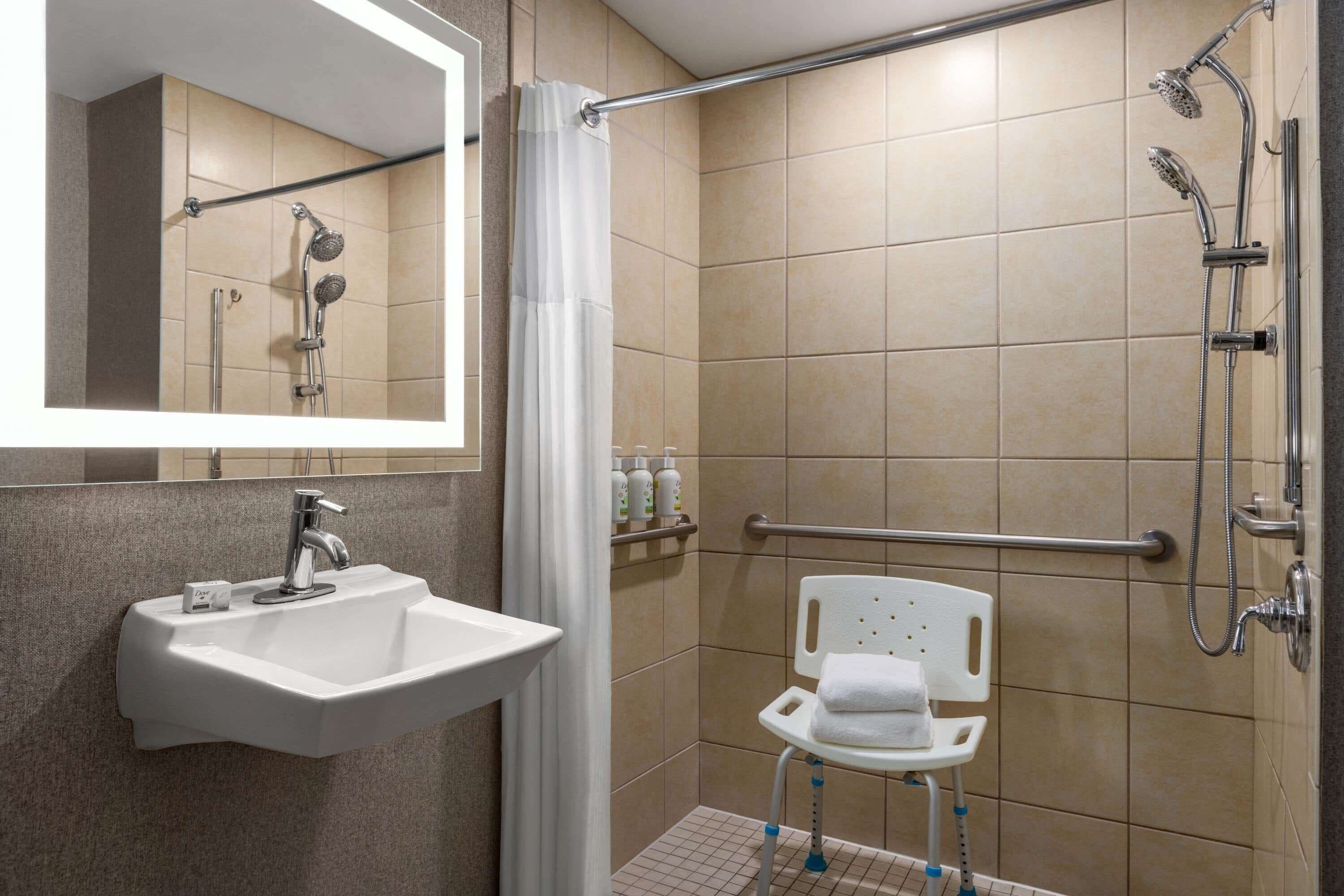 A bathroom with a sink, mirror, shower, and a wheelchair-accessible chair.