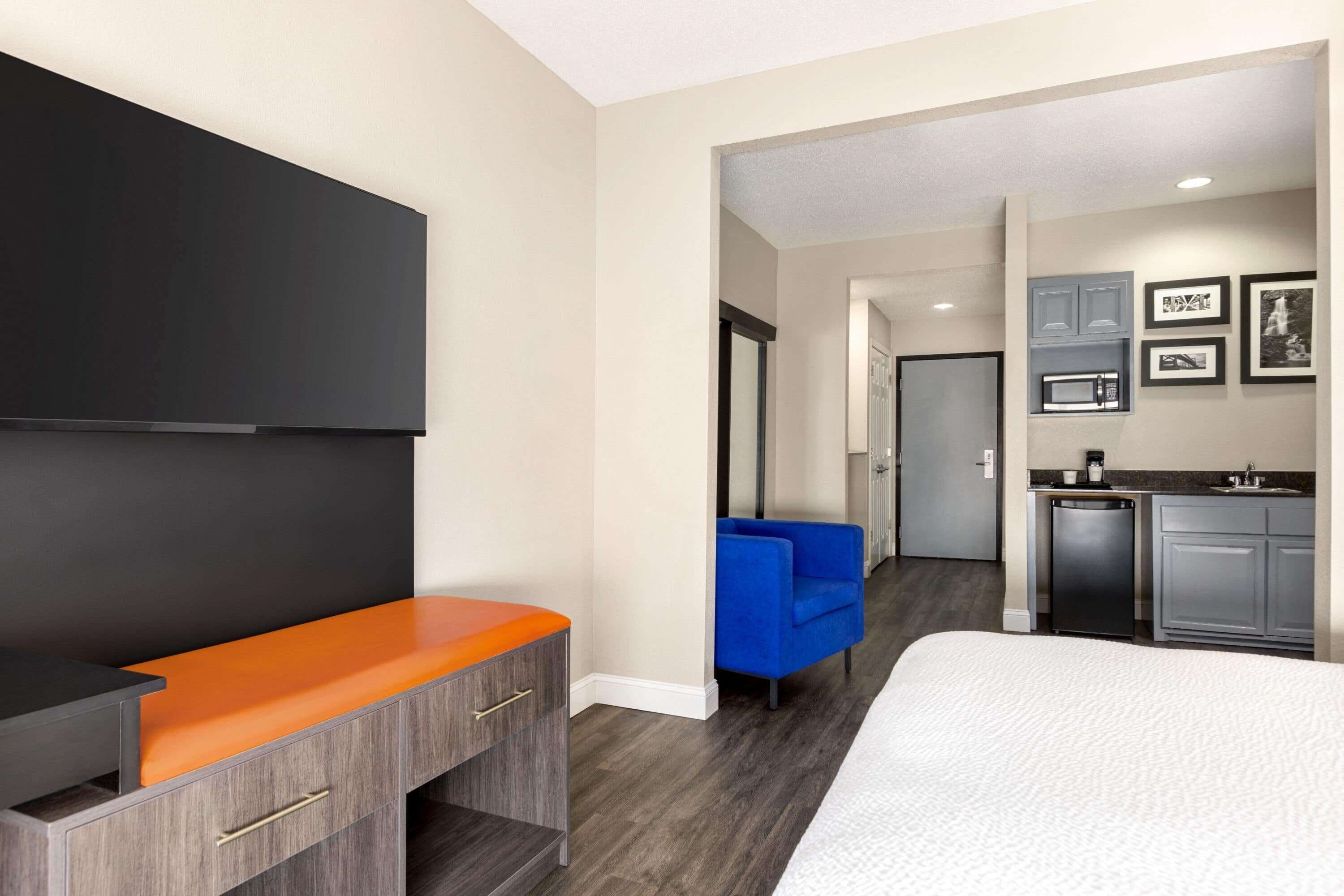 A modern hotel room with a flat-screen TV, a bed, a desk with a laptop, a blue armchair, and a kitchenette.