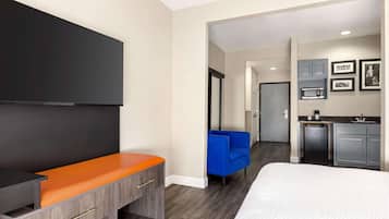 A modern hotel room with a flat-screen TV, a bed, a desk with a laptop, a blue armchair, and a kitchenette.
