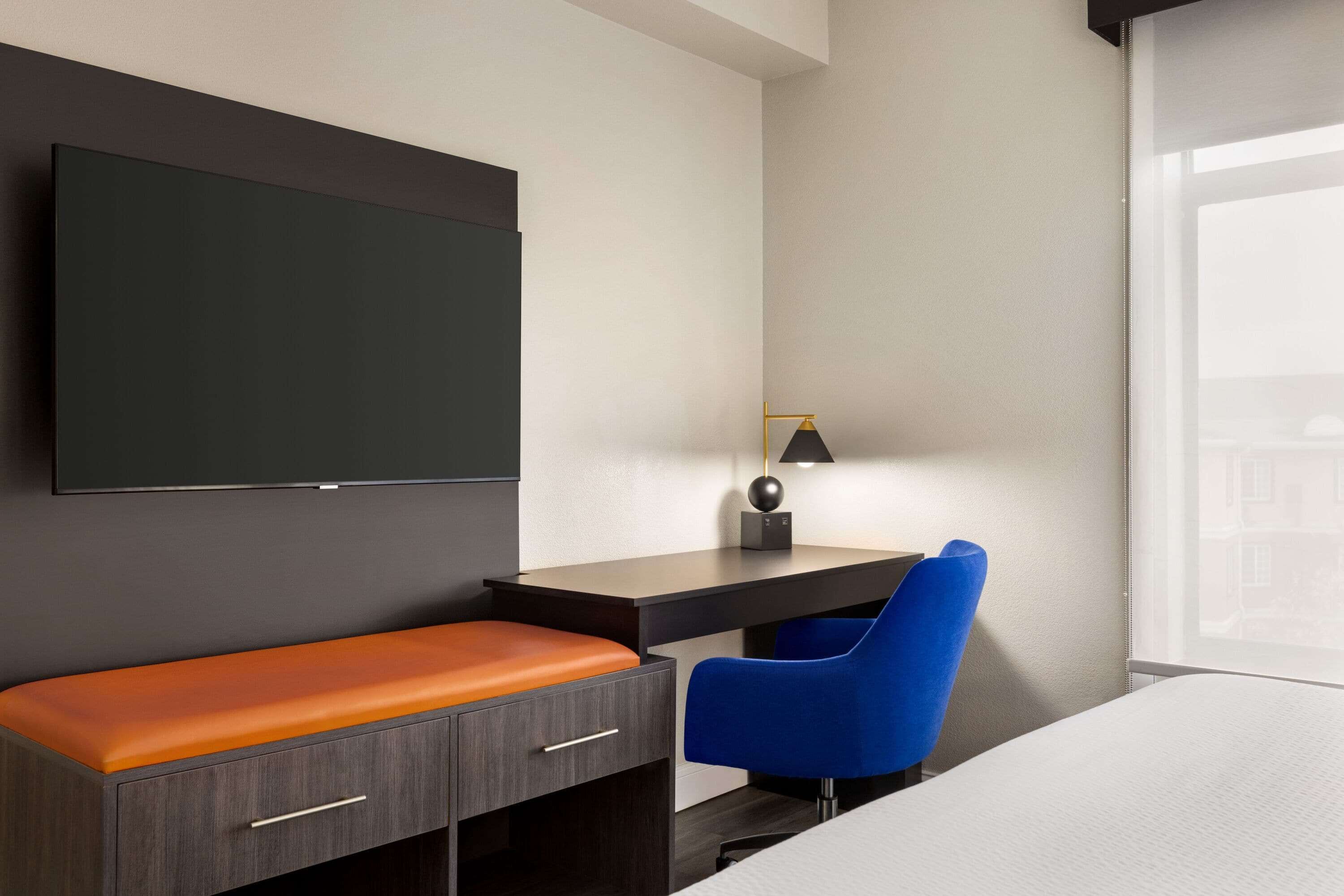 A modern hotel room with a flat-screen TV, a desk with a chair, a bench, and a window with a view.