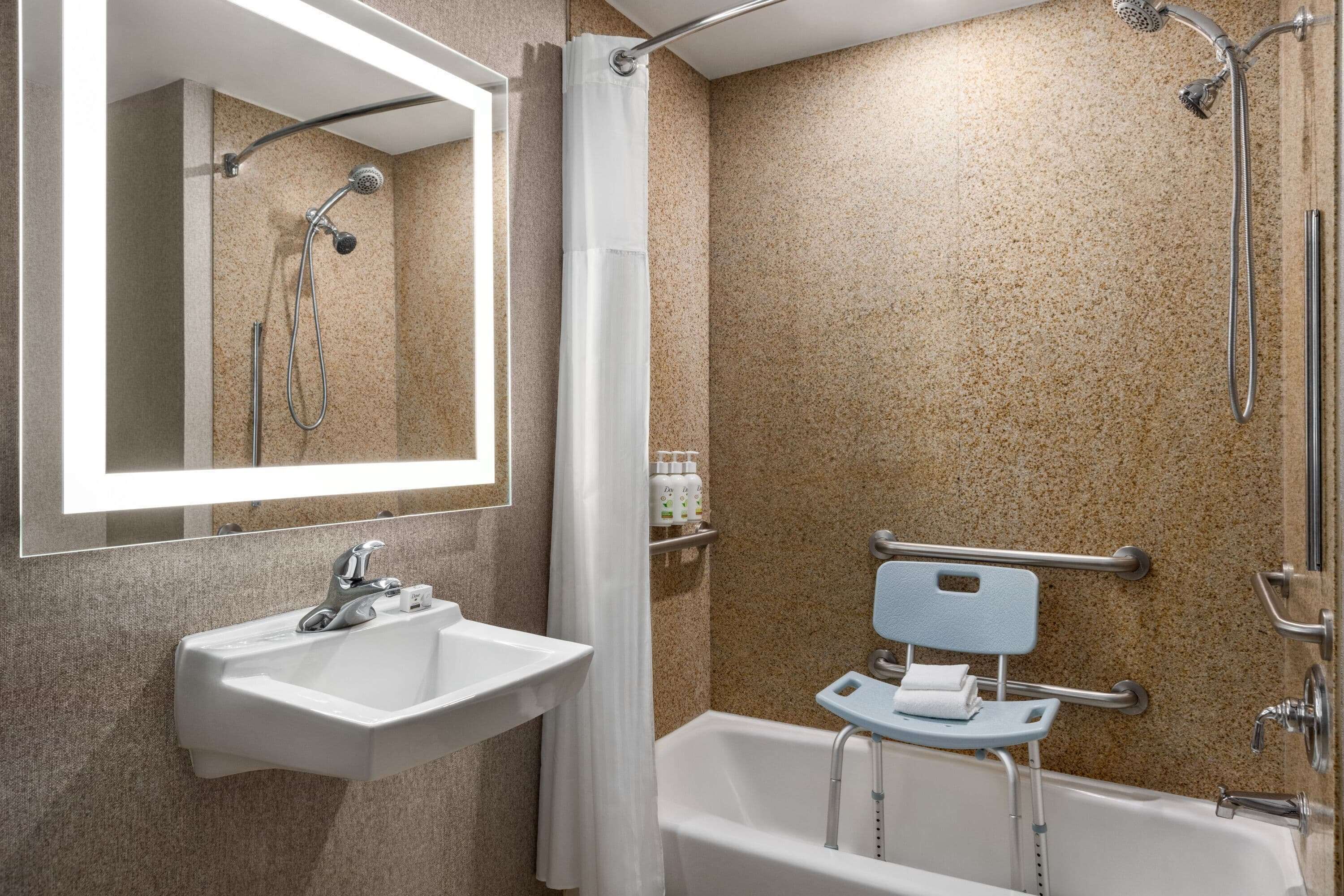 A bathroom with a sink, mirror, shower, and a wheelchair-accessible bench.