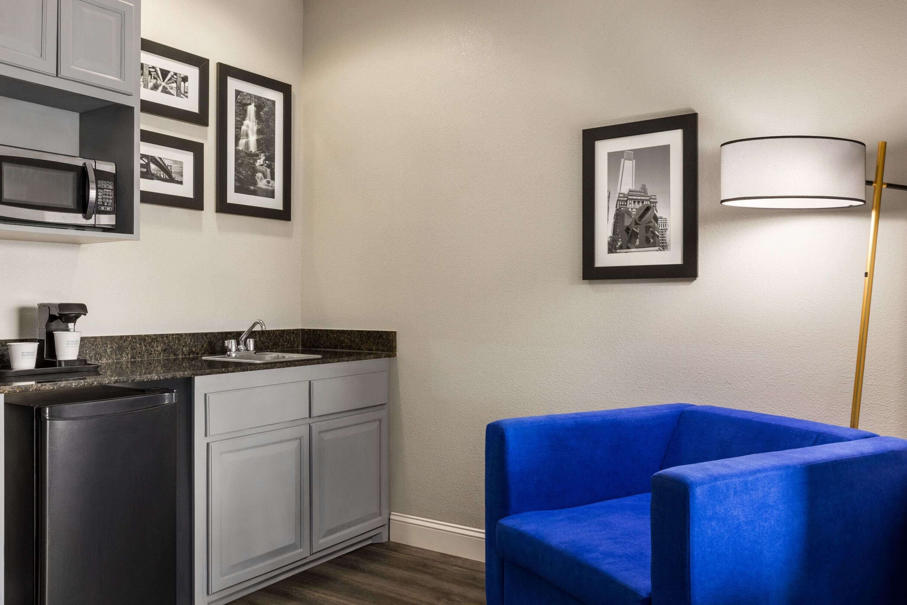 Een moderne keuken met een ingebouwde magnetron, spoelbak en kastjes. Er hangen fotolijstjes aan de muur en er staat een blauwe stoel in de hoek.